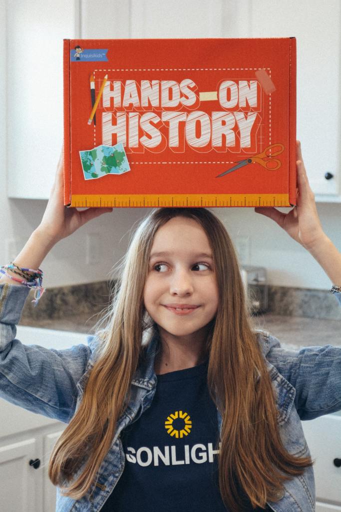 A happy homeschool student, wearing a Sonlight tshirt, holds up a Hands-on History box of history projects.